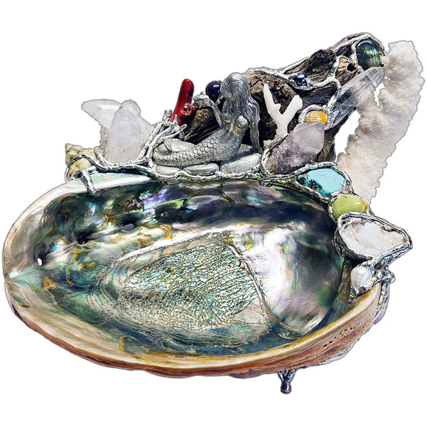 A mermaid rests on a large pink abalone shell, adorned with corals, seashells, gemstones and driftwood