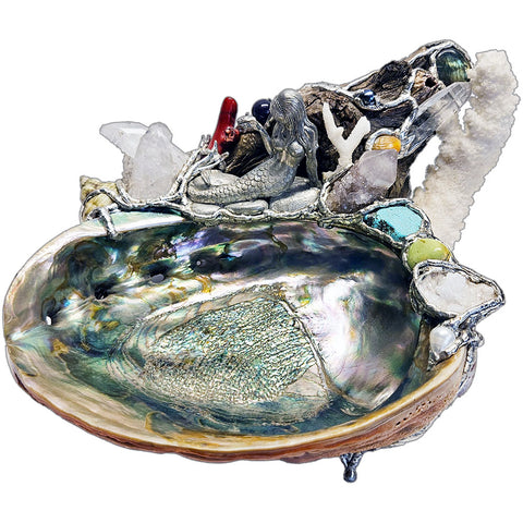 A mermaid rests on a large pink abalone shell, adorned with corals, seashells, gemstones and driftwood